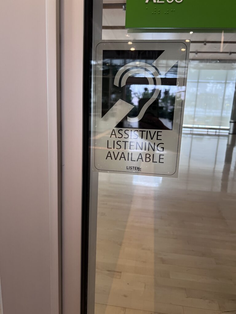 Assistive Listening Available sign on a glass door, below a room number sign with braille