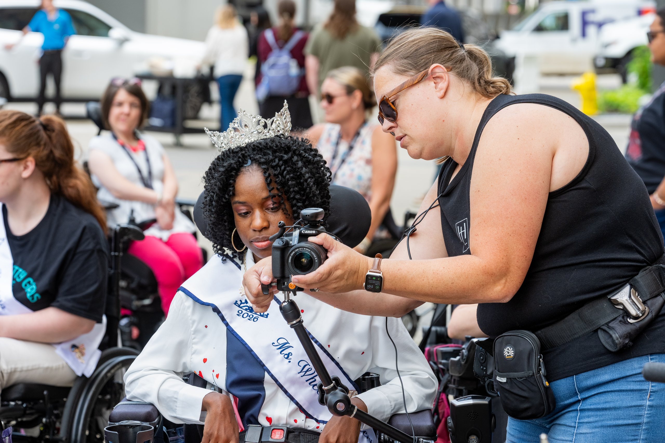 A Hopelynd photographer showing a Ms. Wheelchair America delegate in a power wheelchair how to use a camera during the Walk and Roll in Grand Rapids