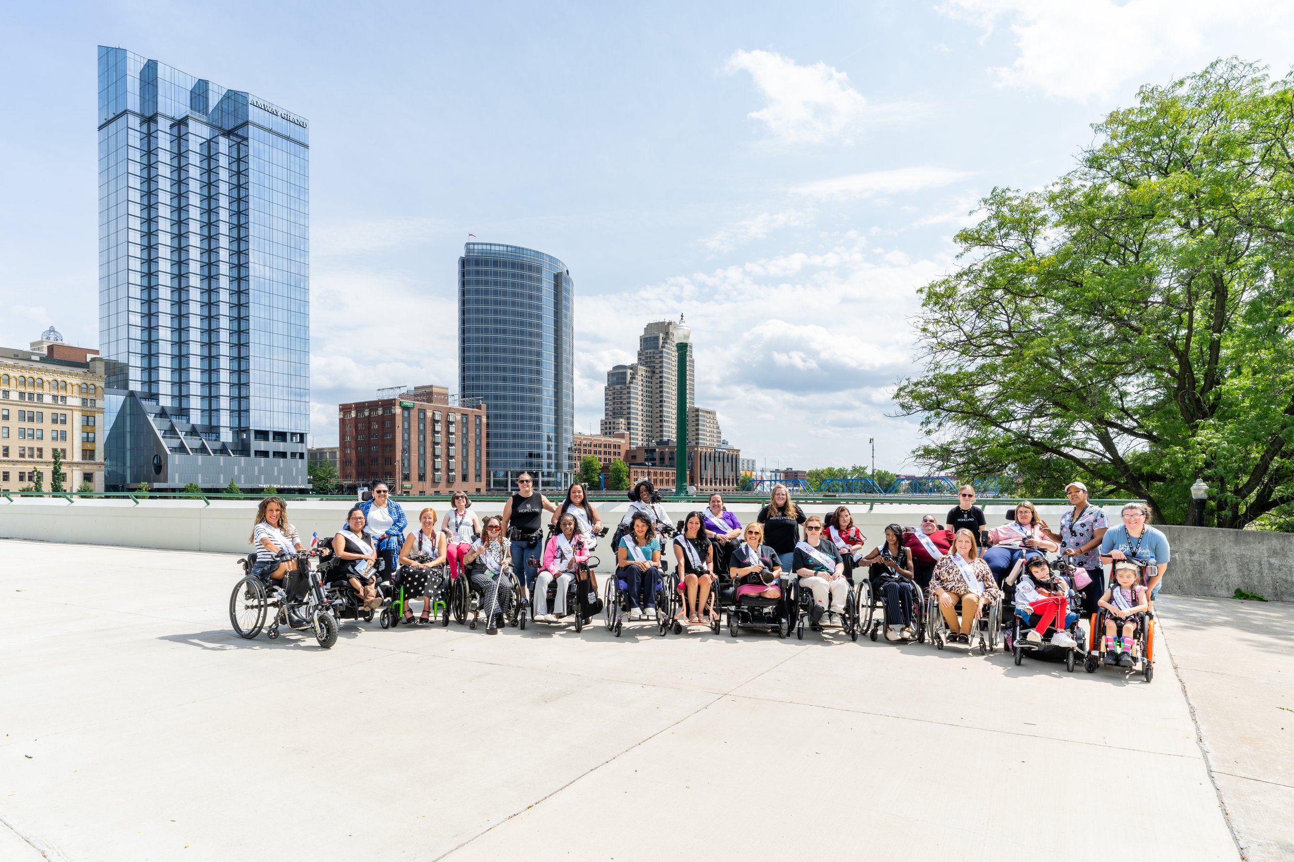 Ms. Wheelchair America 2027 delegates in wheelchairs and powerchairs gathered for a group photo on a Grand Rapids plaza with the downtown skyline behind them during the Hopelynd Walk and Roll