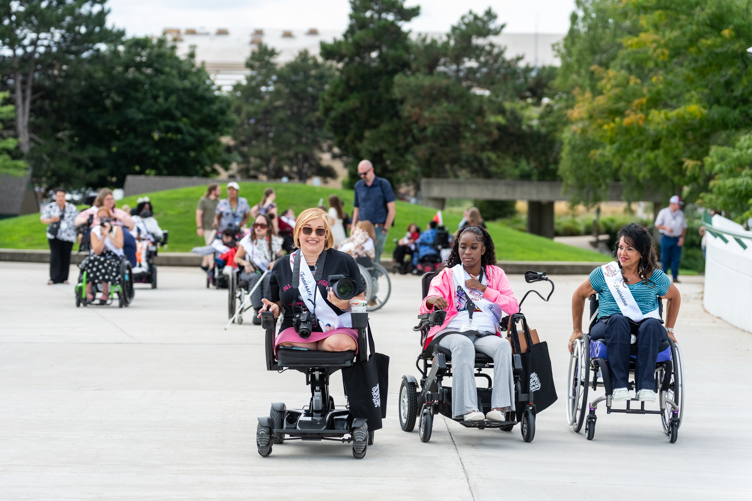 Ms. Wheelchair America delegates in power wheelchairs and manual wheelchairs rolling across a Grand Rapids plaza during the Walk and Roll