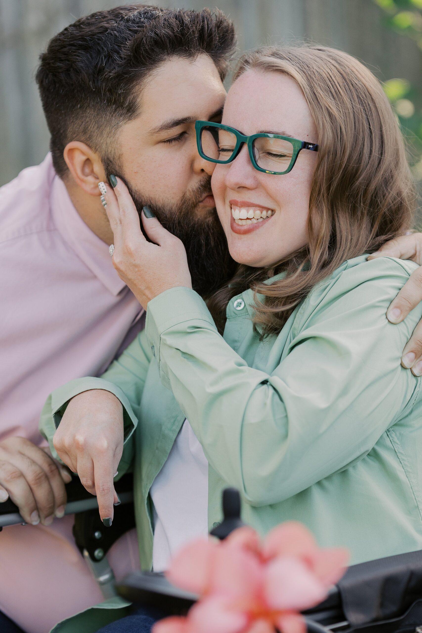 Saleem kissing Katie's cheek as she smiles from her power wheelchair, pink flowers in front
