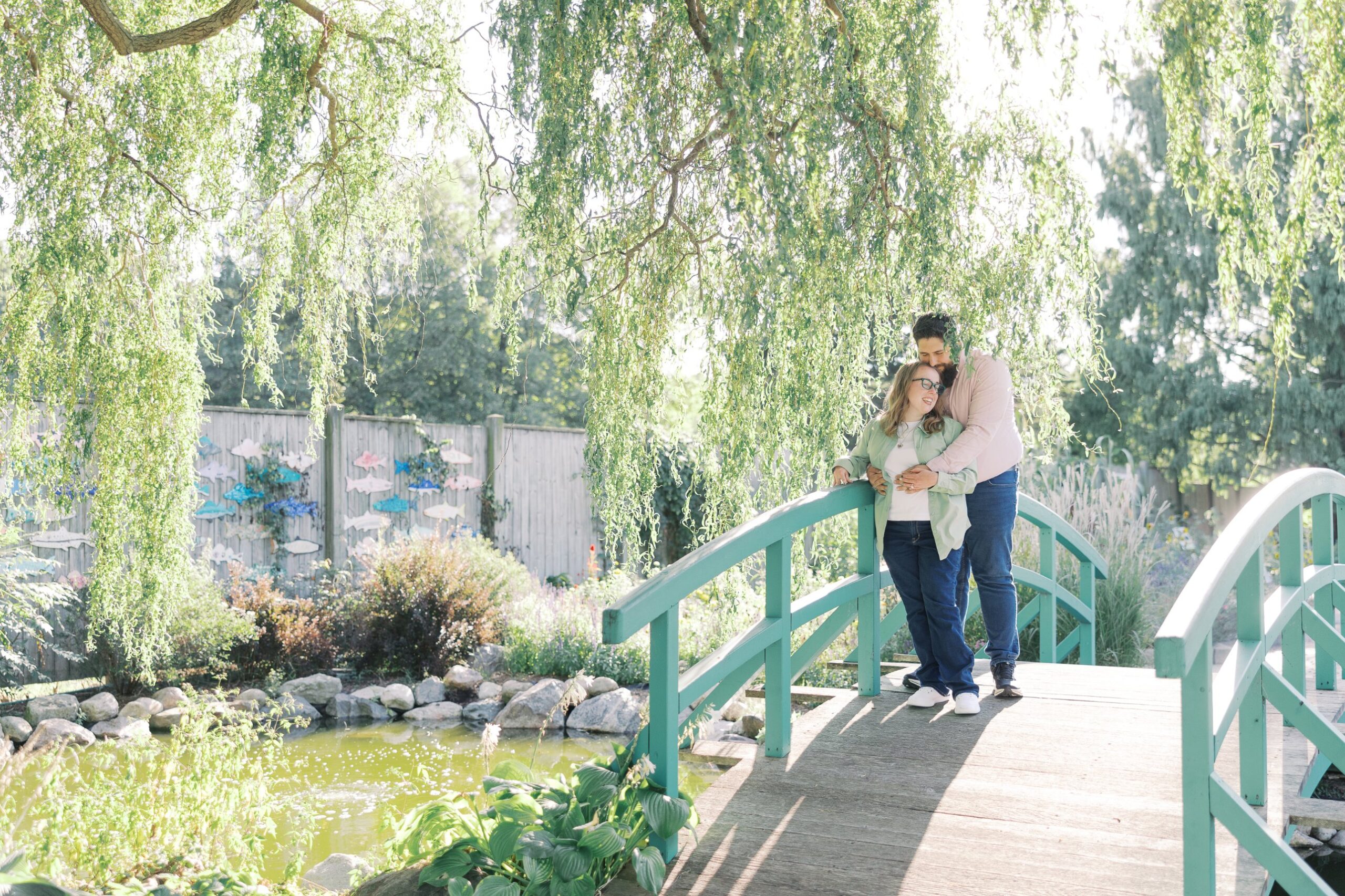 Katie and Saleem embracing on the green garden bridge under a willow tree