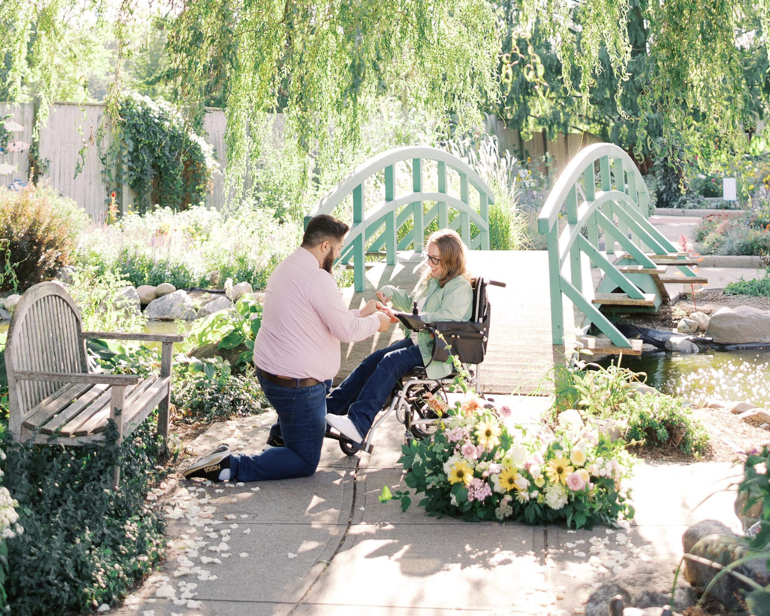 Saleem on one knee proposing to Katie, a powerchair user, as she sits in her power wheelchair beside the green garden bridge at Michigan State University