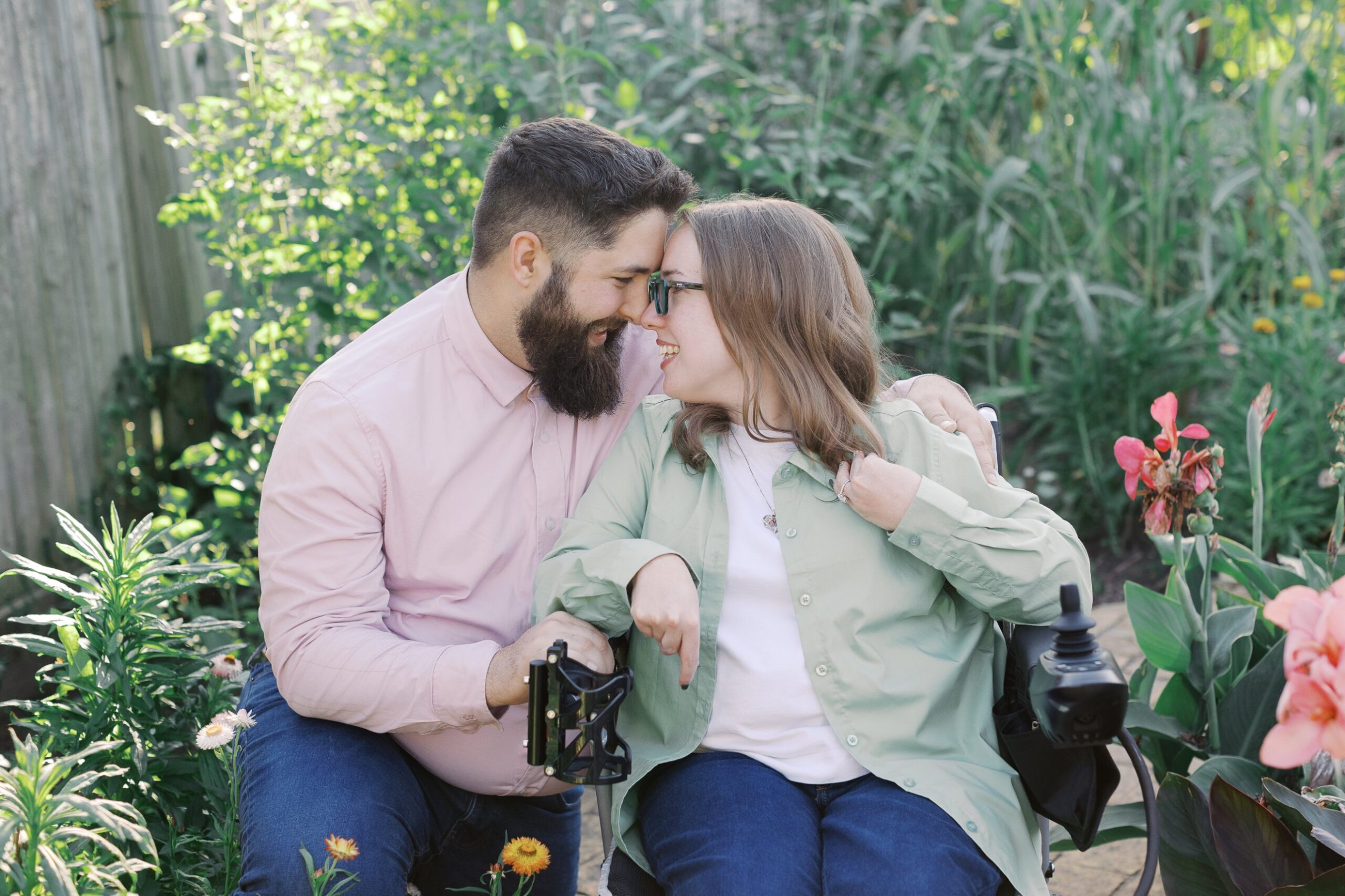 Saleem and Katie touching foreheads and laughing as she sits in her power wheelchair among pink flowers