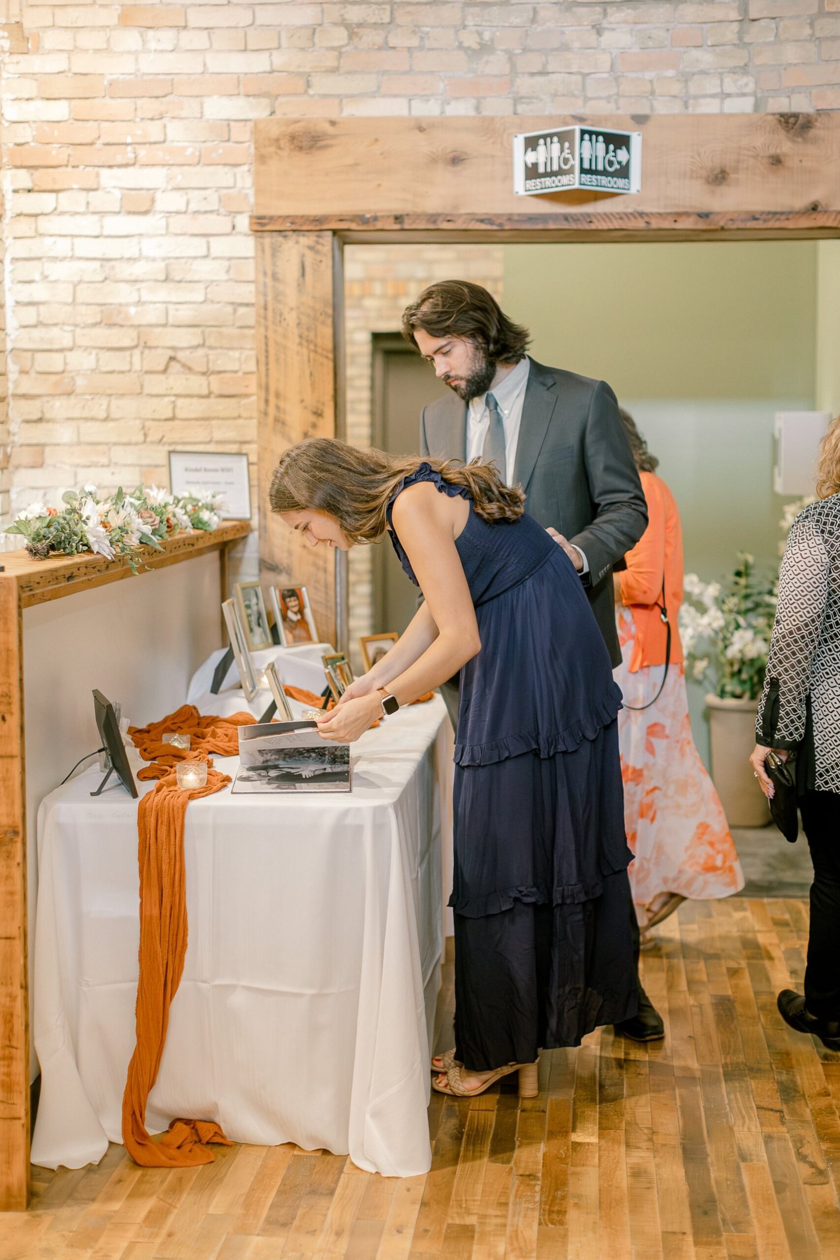 A wide doorway next to the bar in the Kindel Room, with restroom signs showing the accessibility symbol and arrows pointing to restrooms on the left and right. Wedding guests look at photos on a table beside it.