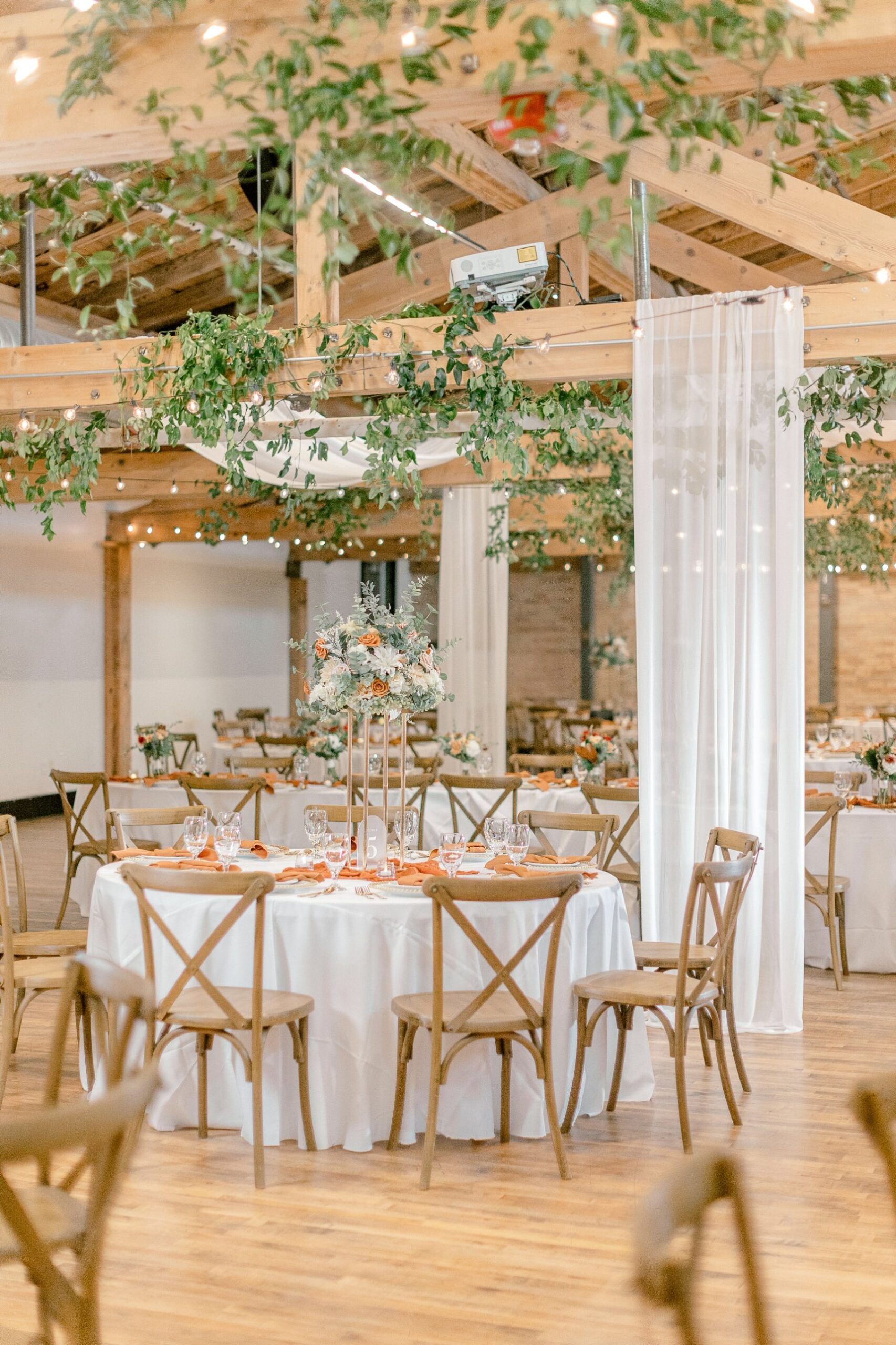 The Kindel Room set for a wedding reception: round tables with white linens and wooden cross-back chairs on a hardwood floor, with greenery and string lights hanging from the wood beams.