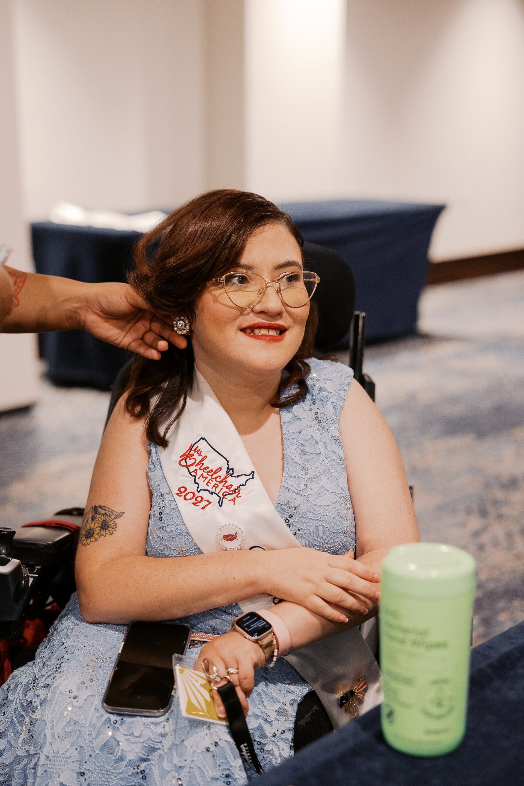 A Ms. Wheelchair America delegate in a light blue gown smiling in her wheelchair while getting her hair done before the crowning ceremony