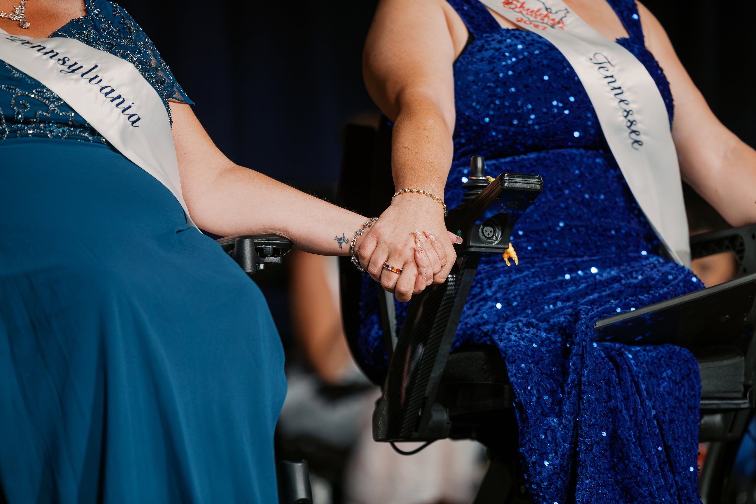 Close-up of Jamie Clendening and Whitney Lavender holding hands from their wheelchairs during the Ms. Wheelchair America 2027 crowning ceremony