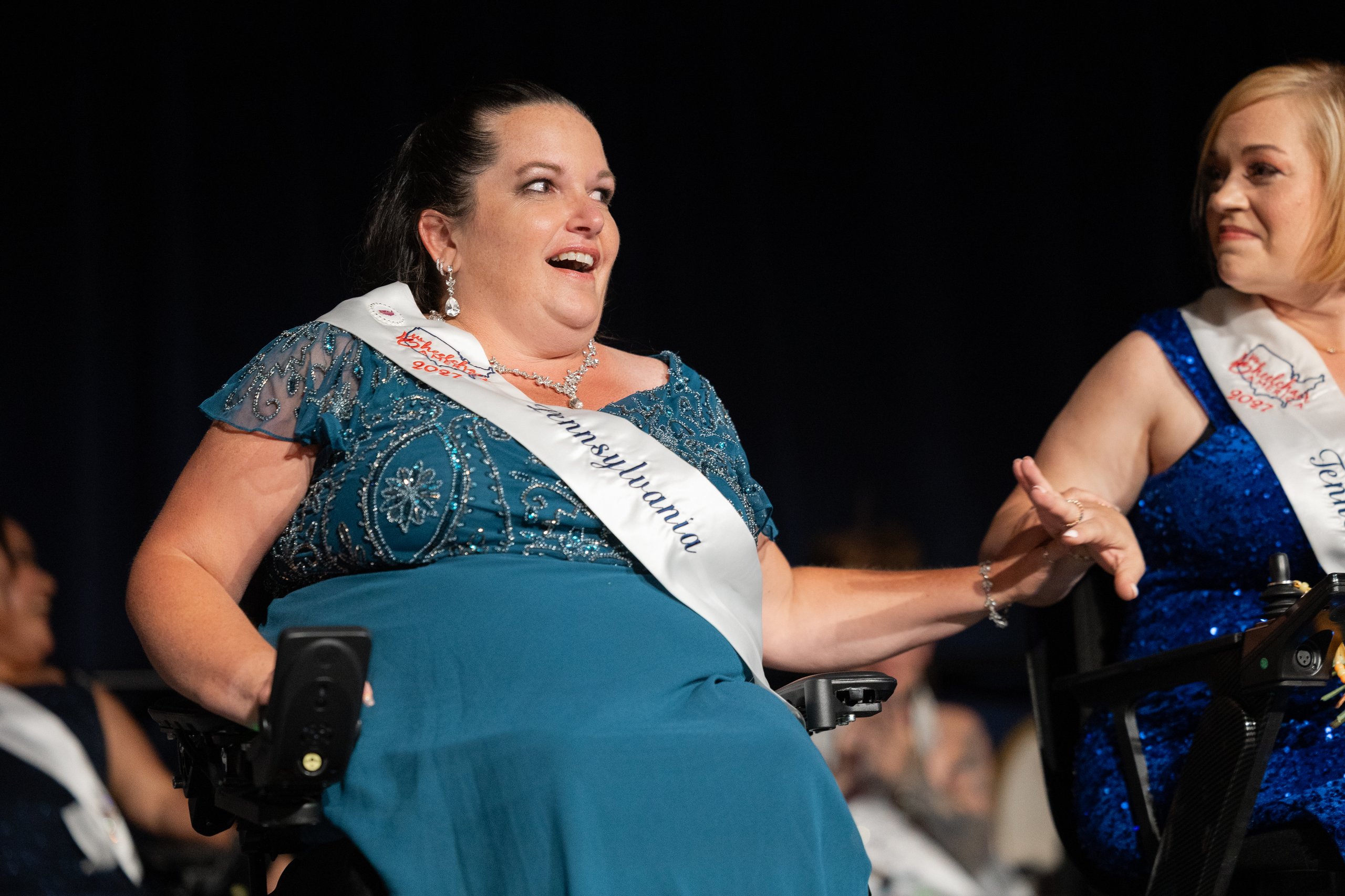 Jamie Clendening of Pennsylvania laughing and holding hands with Whitney Lavender of Tennessee, both wheelchair users, on stage at the Ms. Wheelchair America 2027 crowning ceremony