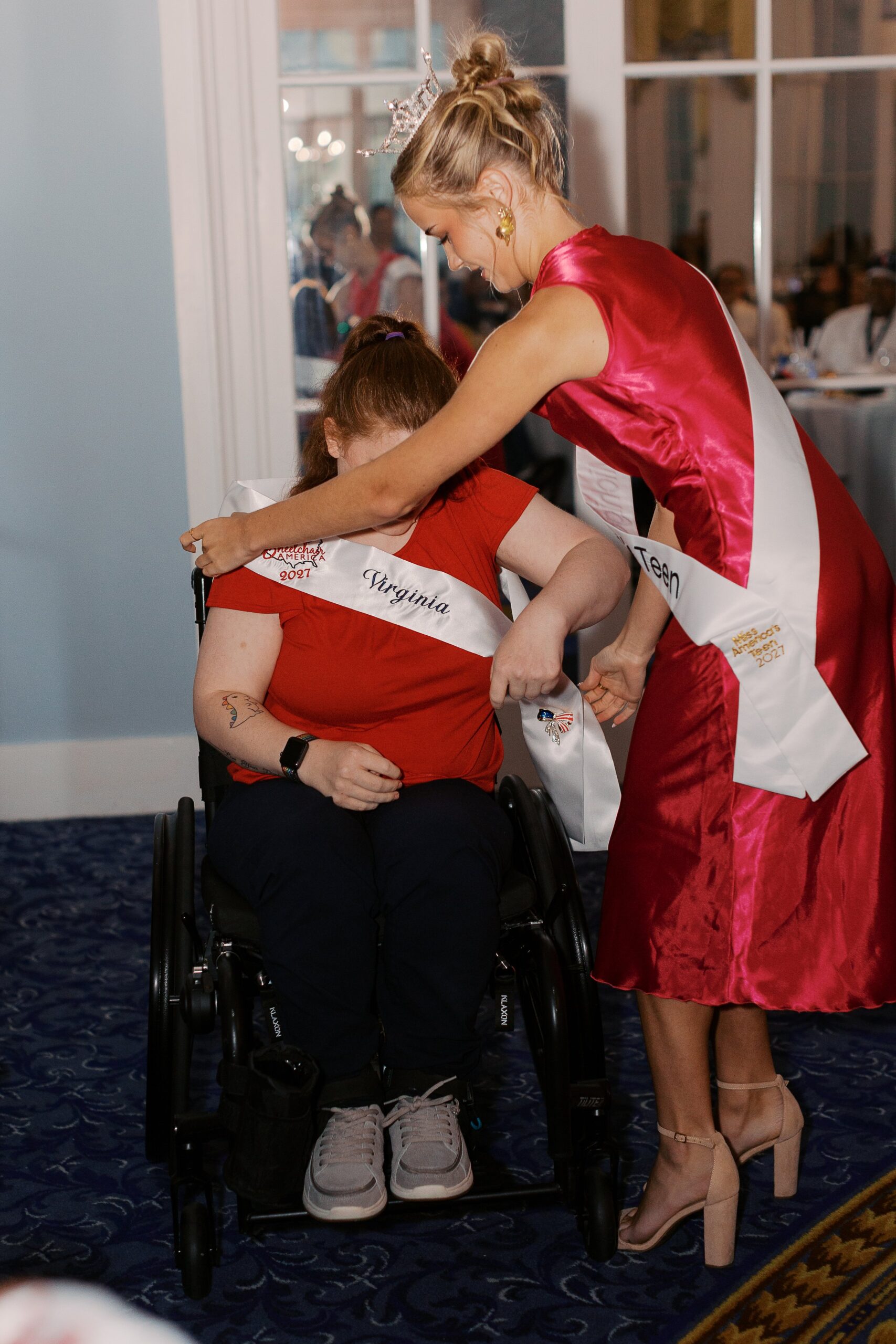 A delegate in a manual wheelchair being re-sashed with a hug during Ms. Wheelchair America Re-Sashing Night