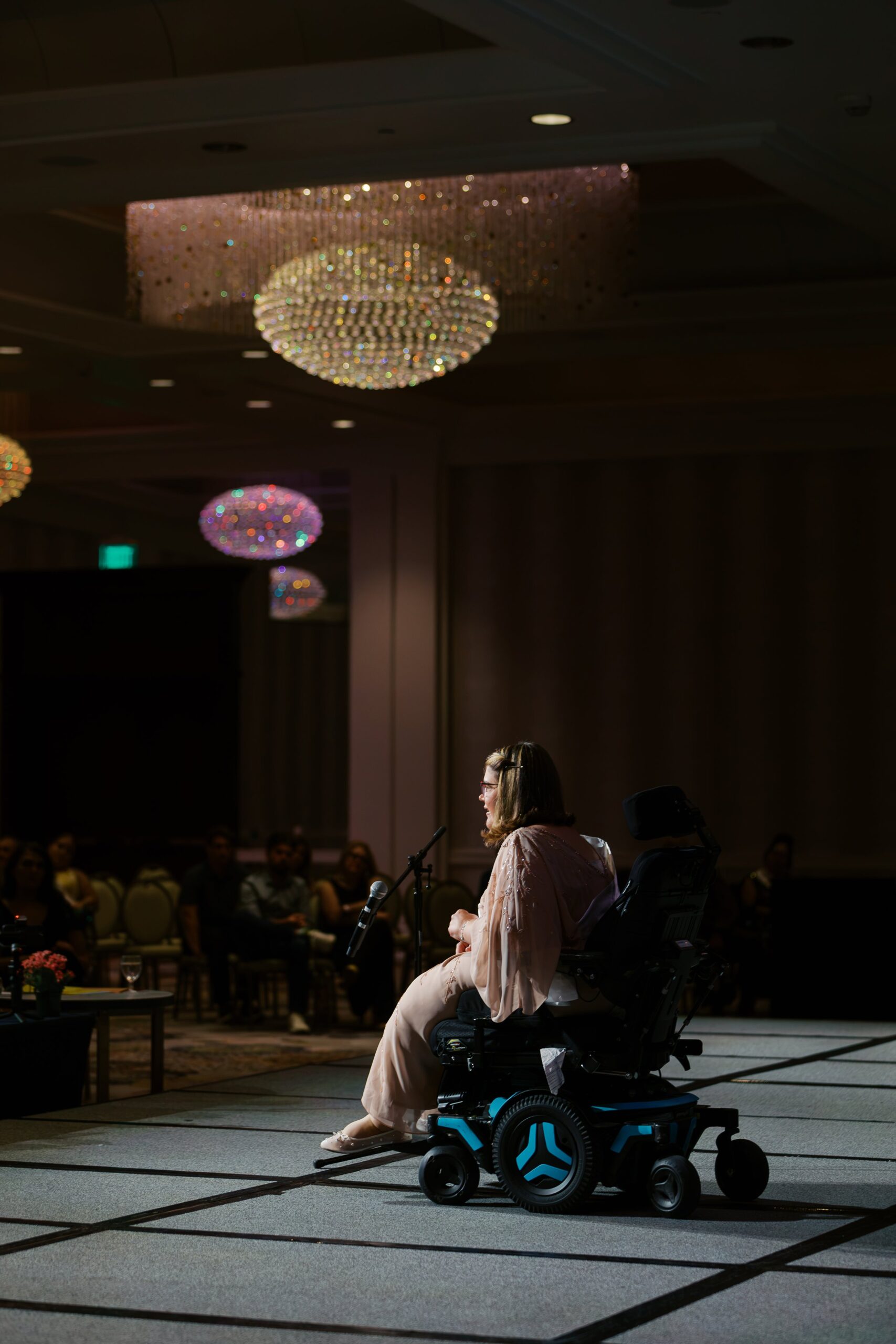 A delegate in a power wheelchair on stage at Ms. Wheelchair America Speech Night under the ballroom chandeliers