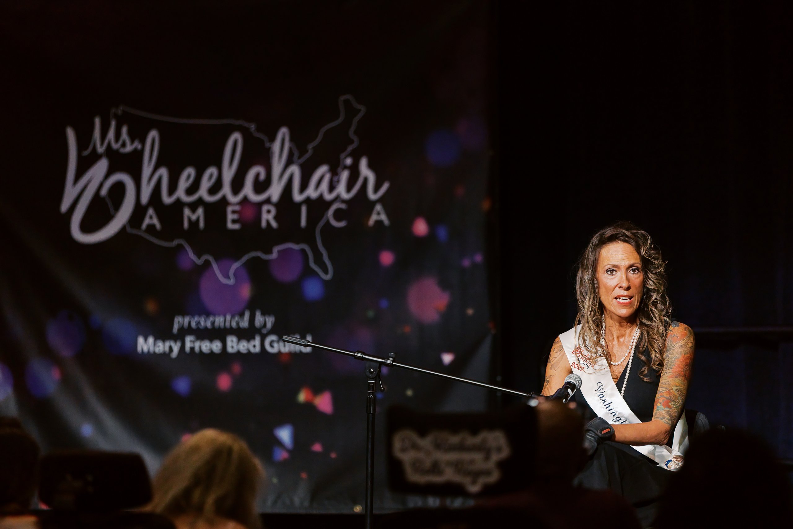 A Ms. Wheelchair America delegate in her wheelchair delivering her platform speech in front of the Ms. Wheelchair America backdrop