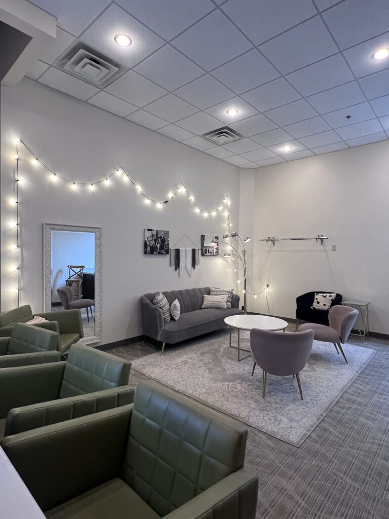 A dressing suite with green and gray seating, a round coffee table, a full-length mirror and string lights.