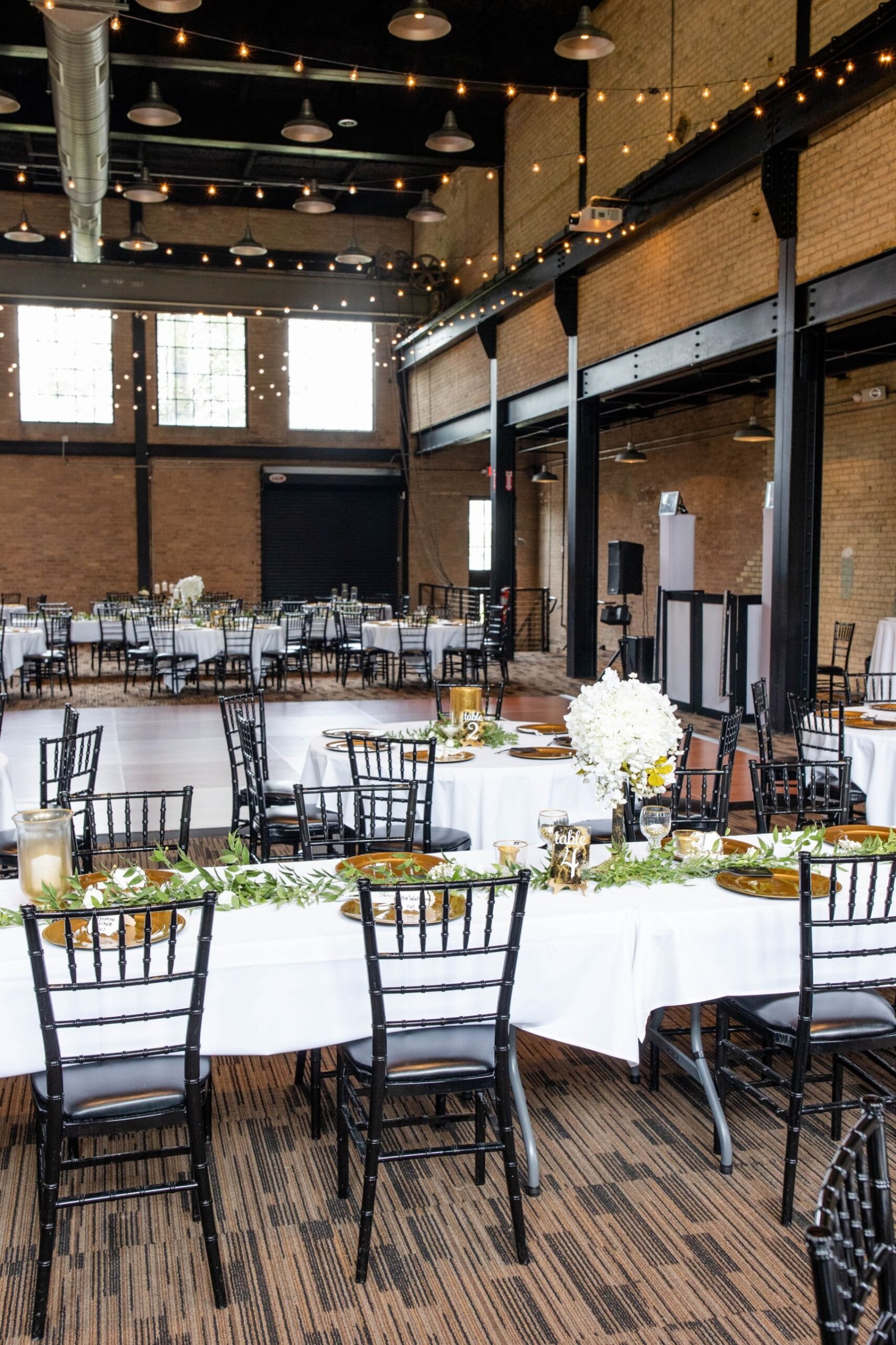 The Great Hall set for a wedding reception, with round tables, black chairs, a dance floor, string lights and tall windows.