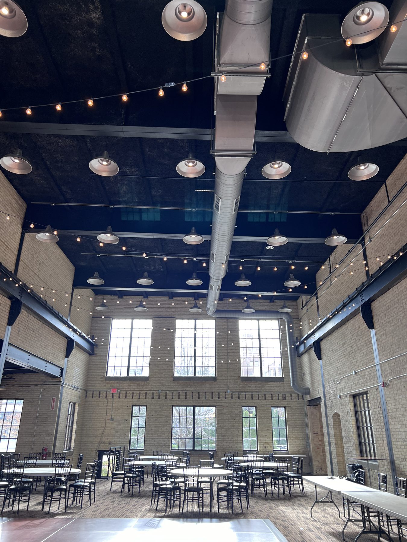 The Great Hall at New Vintage Place, with tall windows, brick walls, string lights and round tables on an open floor.