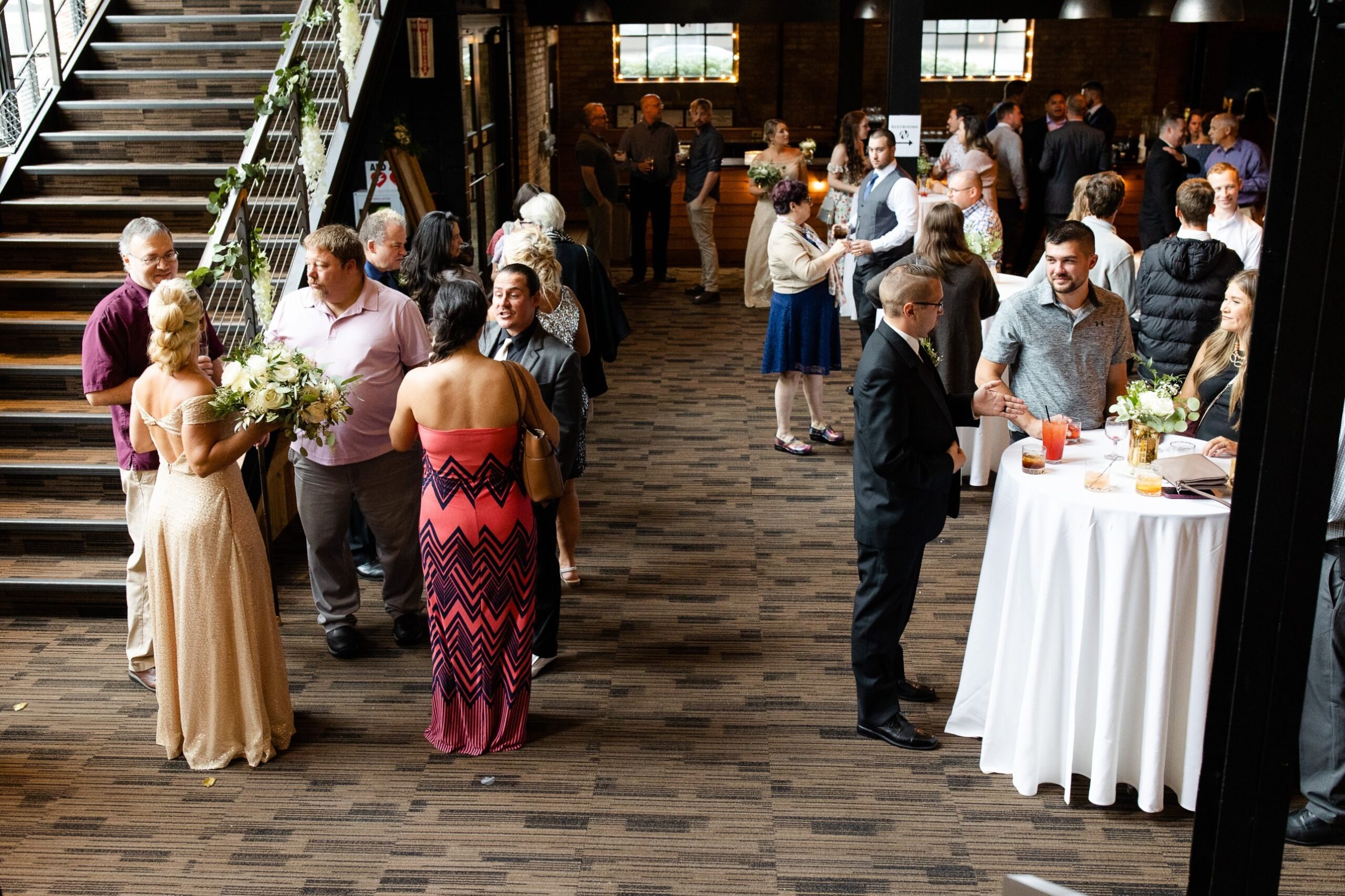 Wedding guests mingle during cocktail hour in the Lounge, with high-top tables on an open carpeted floor, a staircase on the left and a "Restrooms" sign with an arrow near the bar.