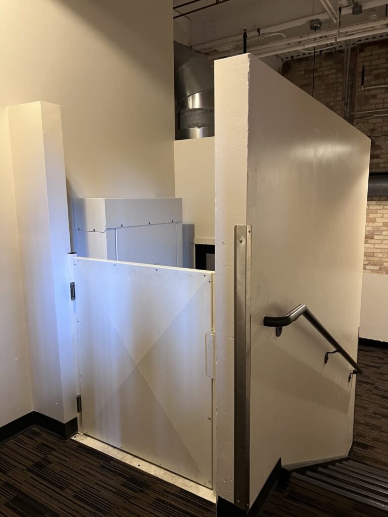 An enclosed wheelchair platform lift with a gate, beside a short set of stairs with a handrail.
