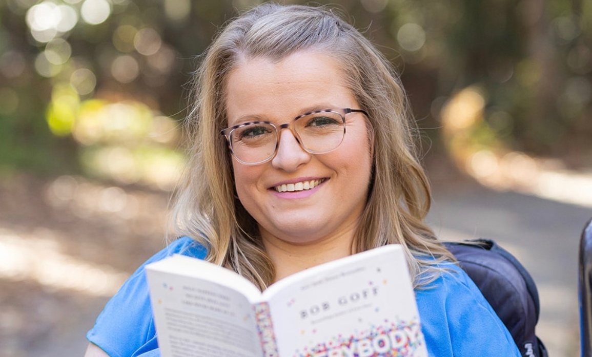 Sara Gaver smiling in her power wheelchair outdoors, holding up the book Everybody Always with a robotic assistive arm.